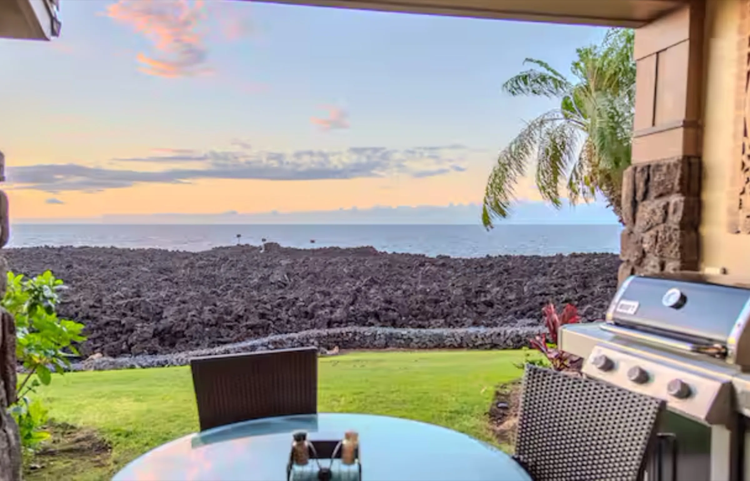 Oceanfront lanai at the Makai Suite with a dining table and grill overlooking the lava-rock shoreline and the Pacific at dusk