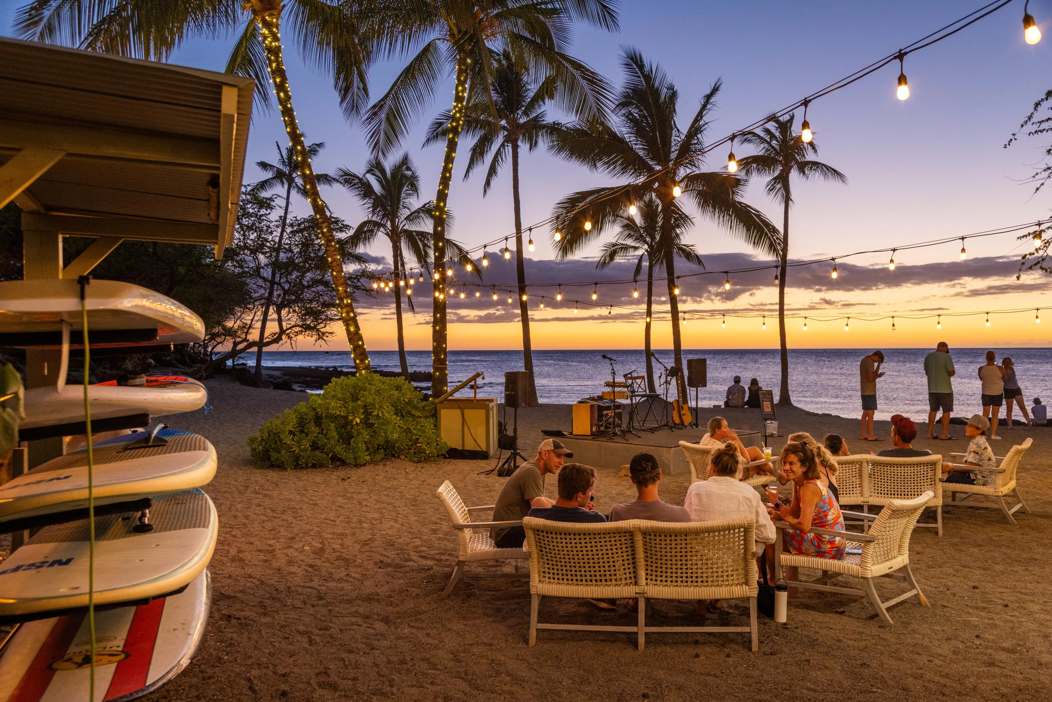 Tables on the sand beneath palms and paddleboards at sunset